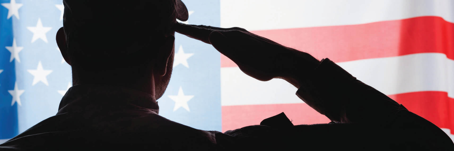 an image from behind of a soldier, in camouflage saluting in front of an image of the American flag