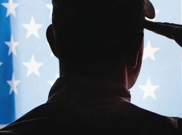 an image from behind of a soldier, in camouflage saluting in front of an image of the American flag