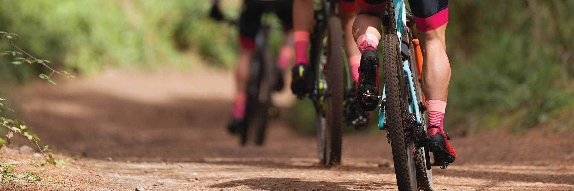 A close-up image of cyclists riding on a dirt trail, focusing on their legs, bike wheels, and pedals