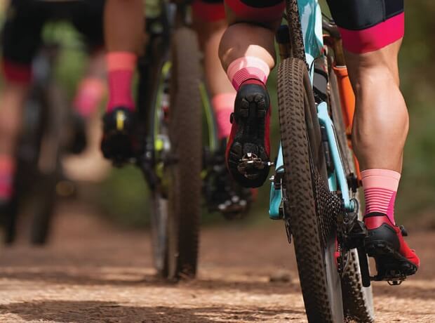 A close-up image of cyclists riding on a dirt trail, focusing on their legs, bike wheels, and pedals