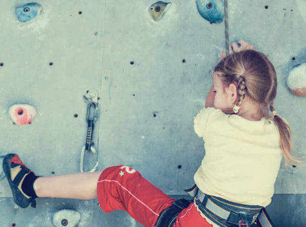 image of a young child with braided hair climbing an indoor rock wall, wearing a harness, red pants, and a light-colored shirt