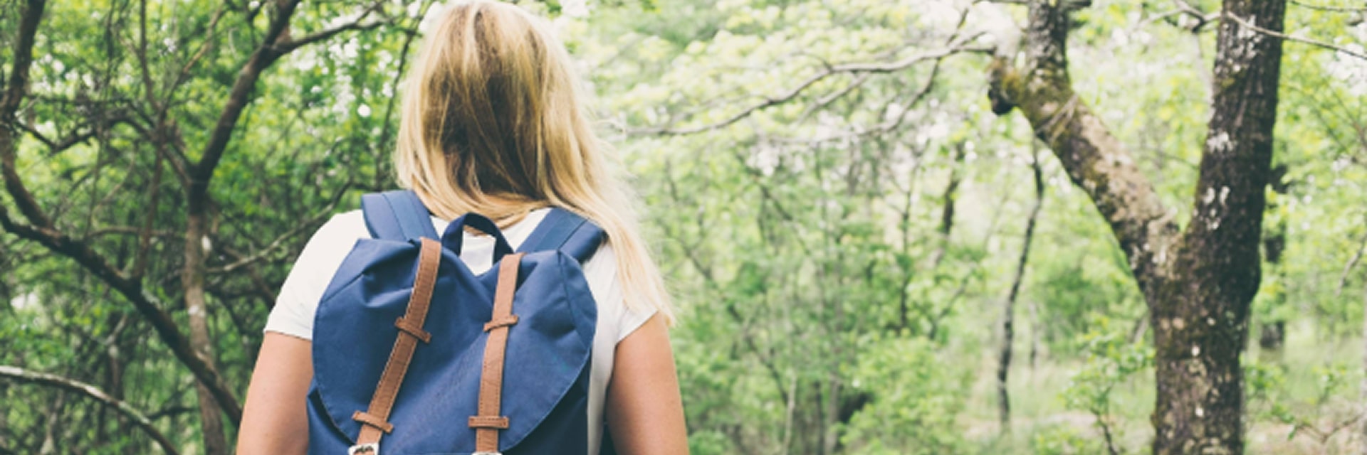 image of a person with long blonde hair hiking on a forest trail, wearing a white shirt, plaid jacket, and a blue Herschel backpack with brown straps