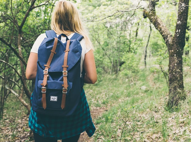 image of a person with long blonde hair hiking on a forest trail, wearing a white shirt, plaid jacket, and a blue Herschel backpack with brown straps