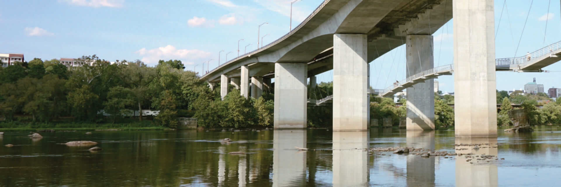 image of large concrete bridge with multiple pillars spanning over a calm river, reflecting on the water, surrounded by green trees under a partly cloudy sky.