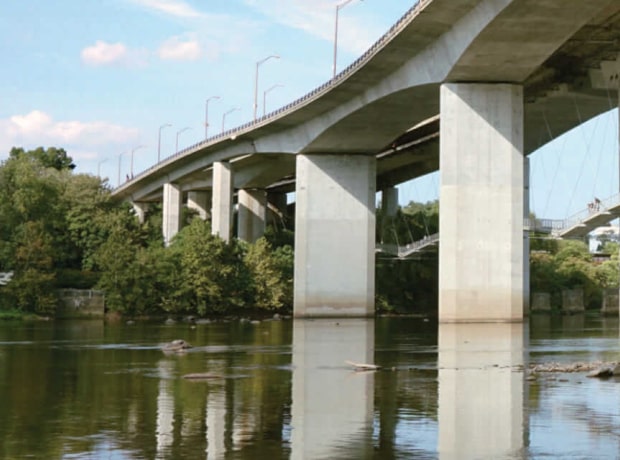 image of large concrete bridge with multiple pillars spanning over a calm river, reflecting on the water, surrounded by green trees under a partly cloudy sky.