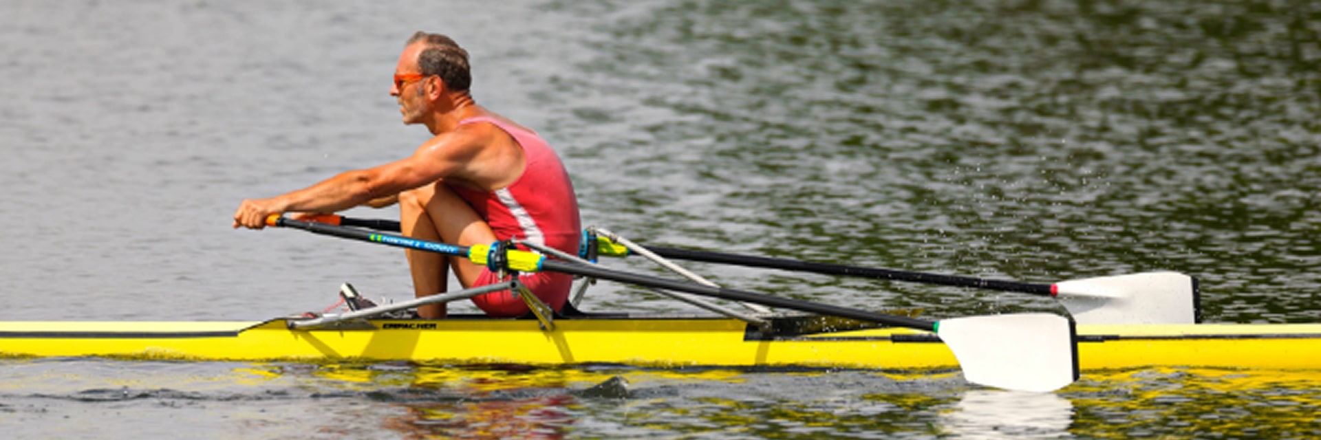 image of a rower in a red uniform rowing a yellow scull on a calm body of water, holding two oars.