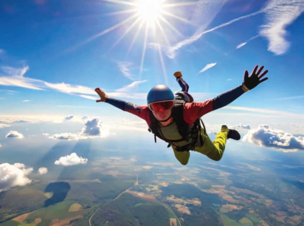 image of a skydiver in a helmet and colorful jumpsuit soaring through the sky with arms and legs spread, above a landscape of fields and forests, under a bright sun with scattered clouds