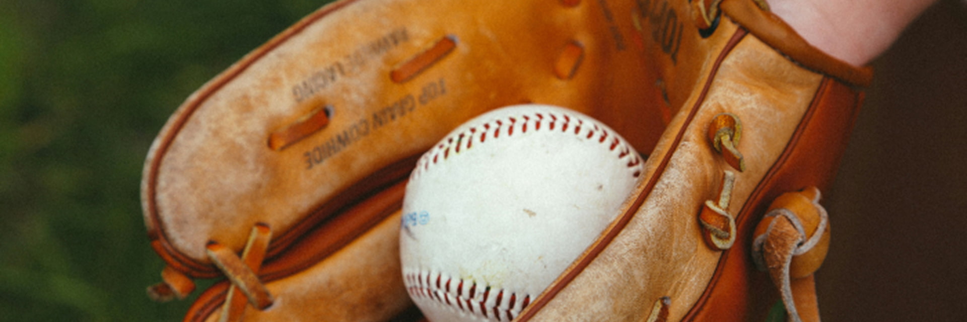 image of a worn leather baseball glove holding a white baseball with red stitching, set against a blurred green field background