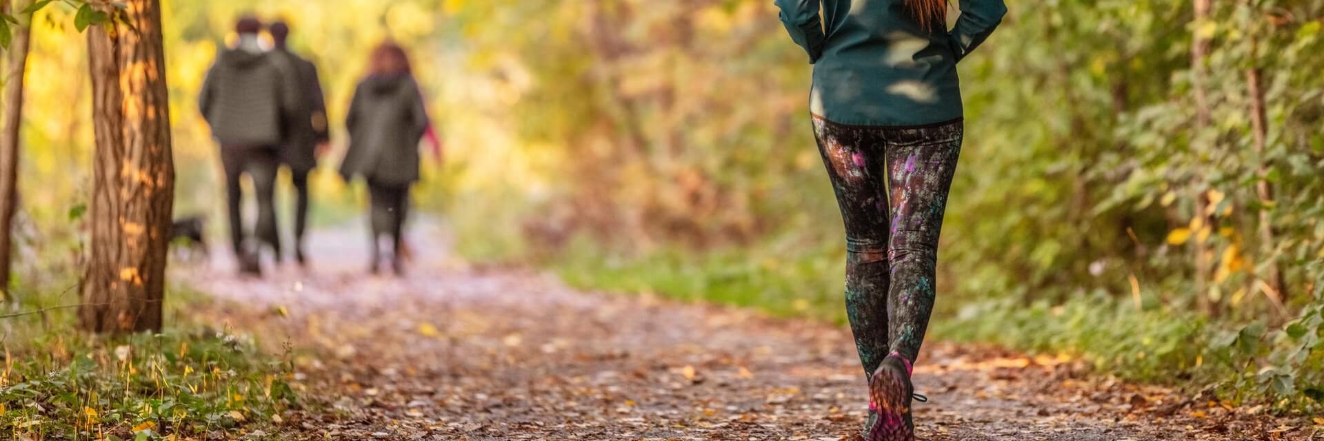 image of a person standing on a dirt path in a forest, facing away from the camera, wearing a green jacket and colorful patterned leggings. In the background, three other people are walking together